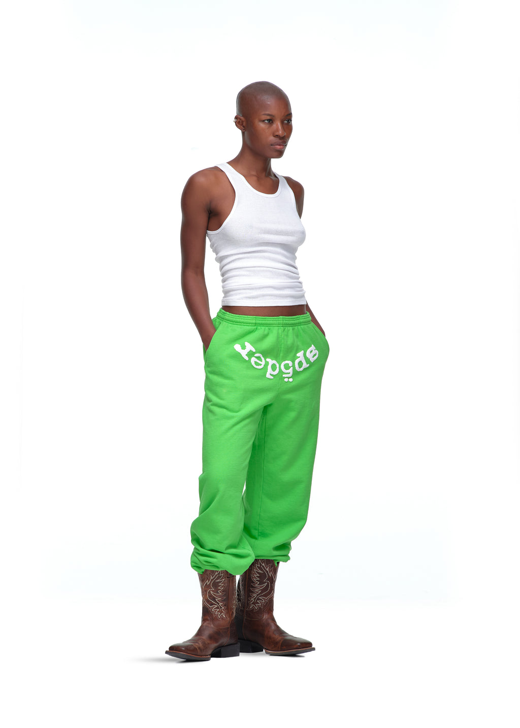 Model wearing bright green LEGACY SWEATPANTS with white "spider" puff print, white tank top, and brown cowboy boots.