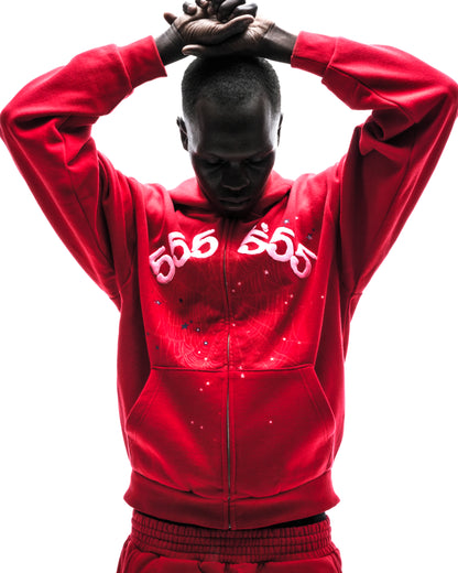 Model in red ANGEL NUMBER ZIP HOODIE with "555" puff print, scattered stars, and zip closure.