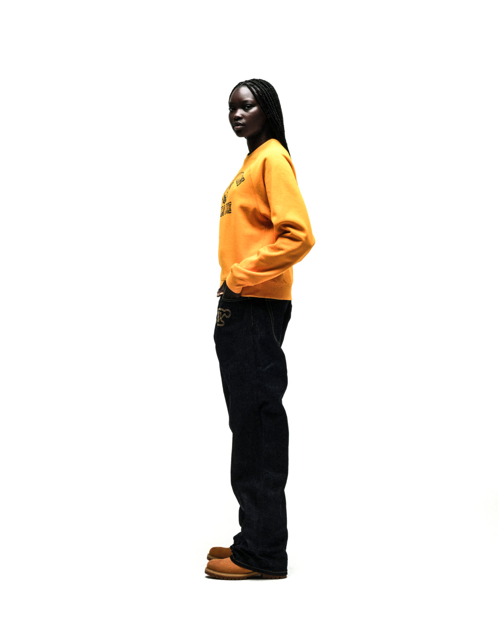 Model wearing a mustard yellow 100% cotton raglan crewneck with a black graphic print, ribbed cuffs, and dark denim jeans.