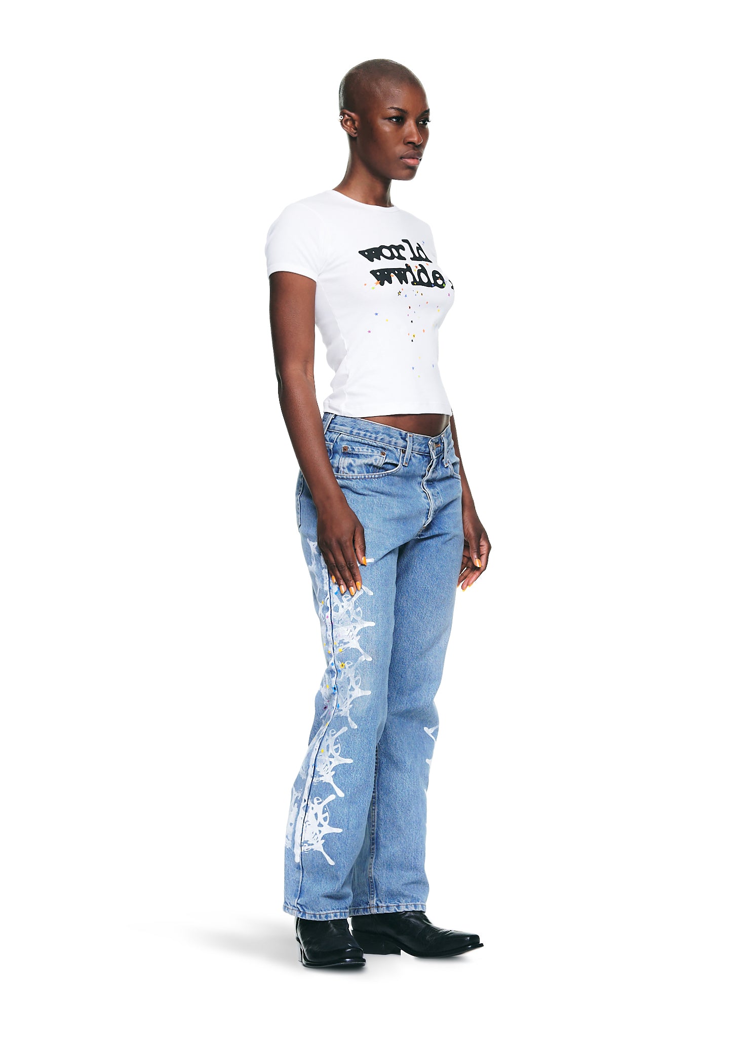 Female model in white 100% cotton "worldwide" graphic baby tee, light wash jeans with white designs, and black boots.