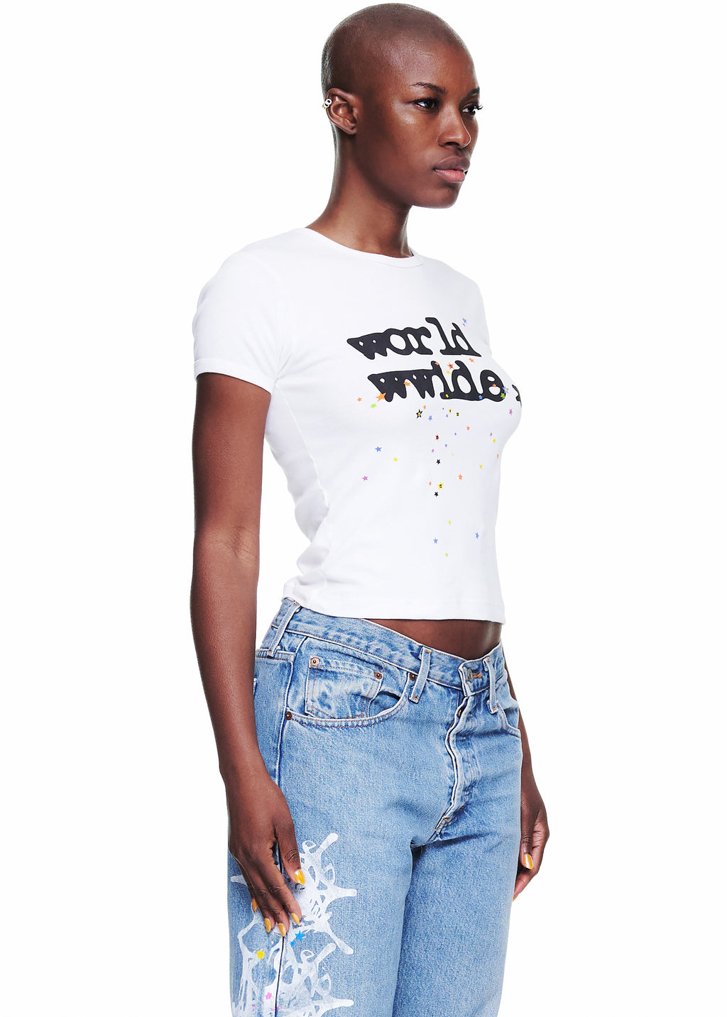 Female model in a white 100% cotton cropped tee with "worldwide" graphic and colorful stars, paired with light wash jeans.