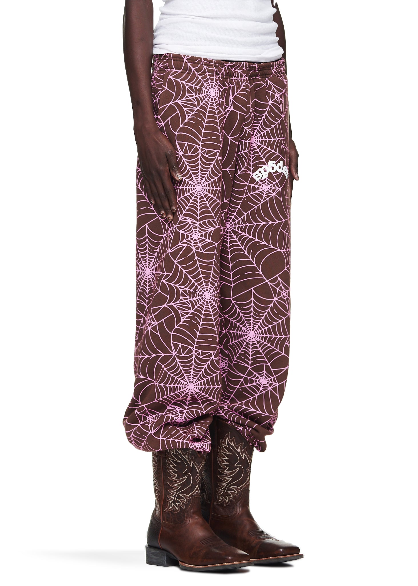 Brown fleece sweatpants with all-over pink cracked web print and "SP5DER" text, worn with cowboy boots.