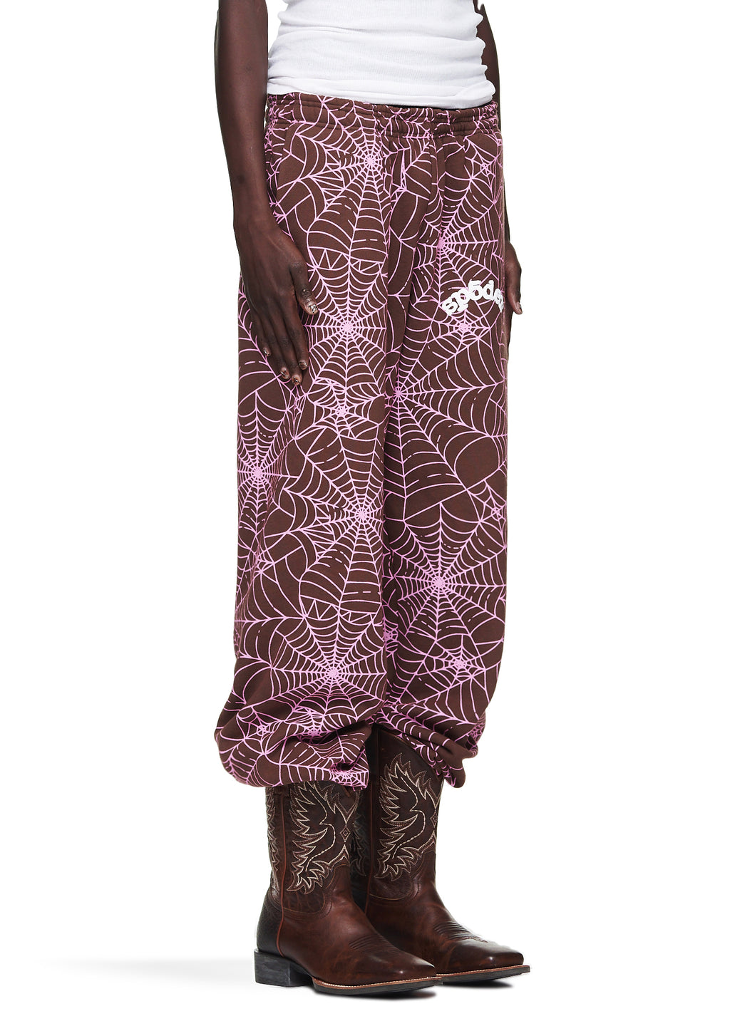 Brown fleece sweatpants with all-over pink cracked web print and "SP5DER" text, worn with cowboy boots.