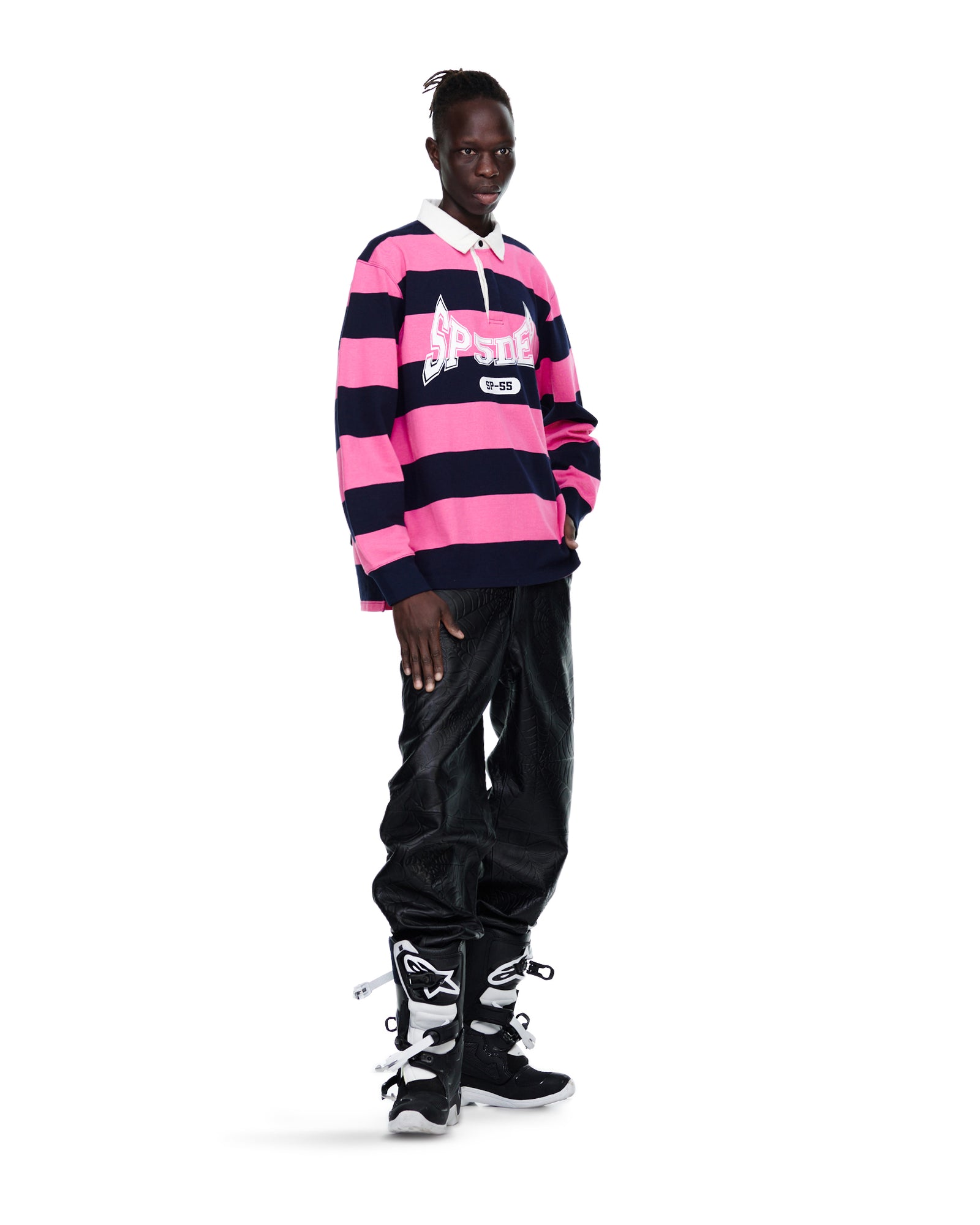 Male model in pink and navy striped T&F PATCH RUGBY SHIRT with 'SP5DER' text, black pants, and boots.