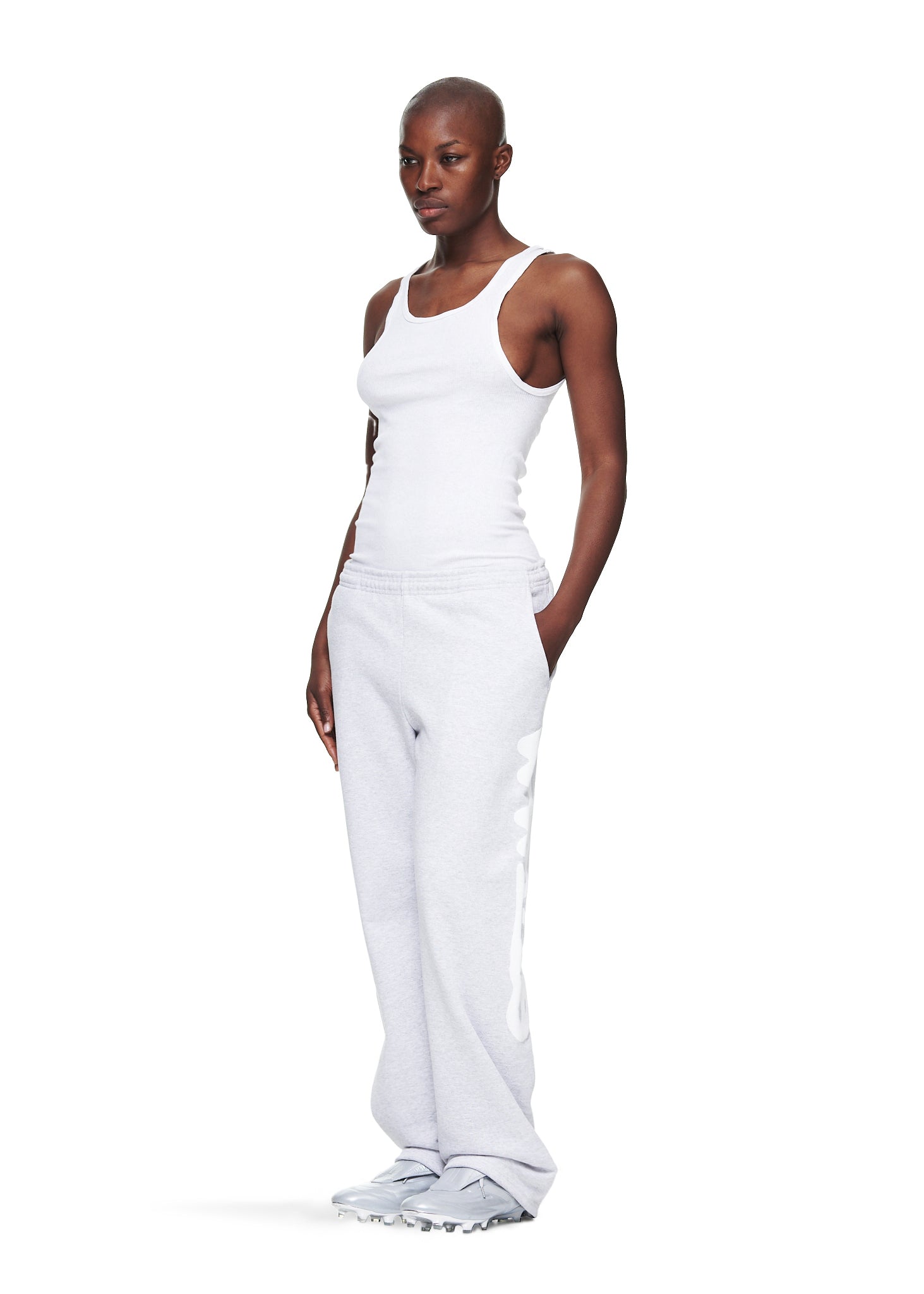 Woman in light grey straight leg sweatpants with "Worldwide" puff print, white tank top, and grey shoes.