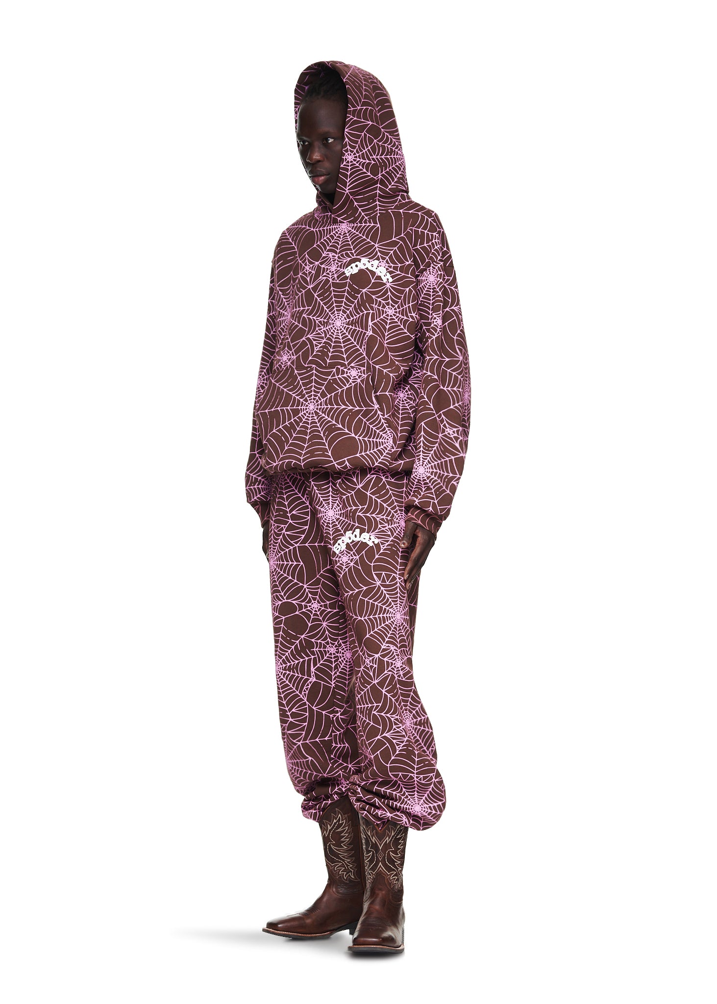 Male model in a brown AOP hoodie and sweatpants with pink cracked web print, wearing cowboy boots.