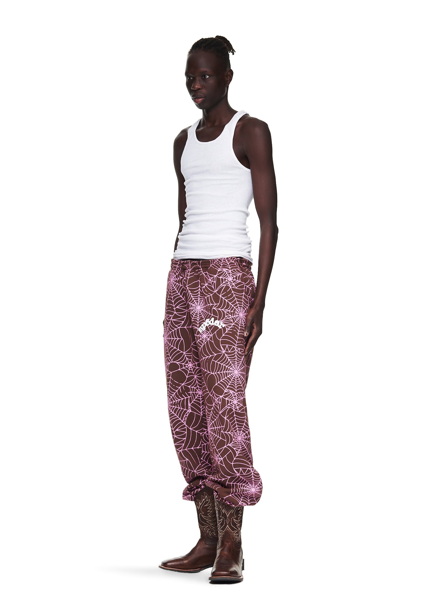Male model in a white tank top and brown AOP SP5DER sweatpants with pink cracked web print and "SP5DER" text.