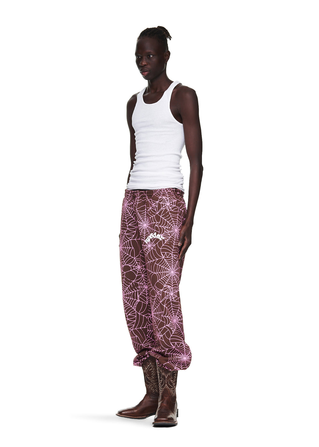 Male model in a white tank top and brown AOP SP5DER sweatpants with pink cracked web print and "SP5DER" text.