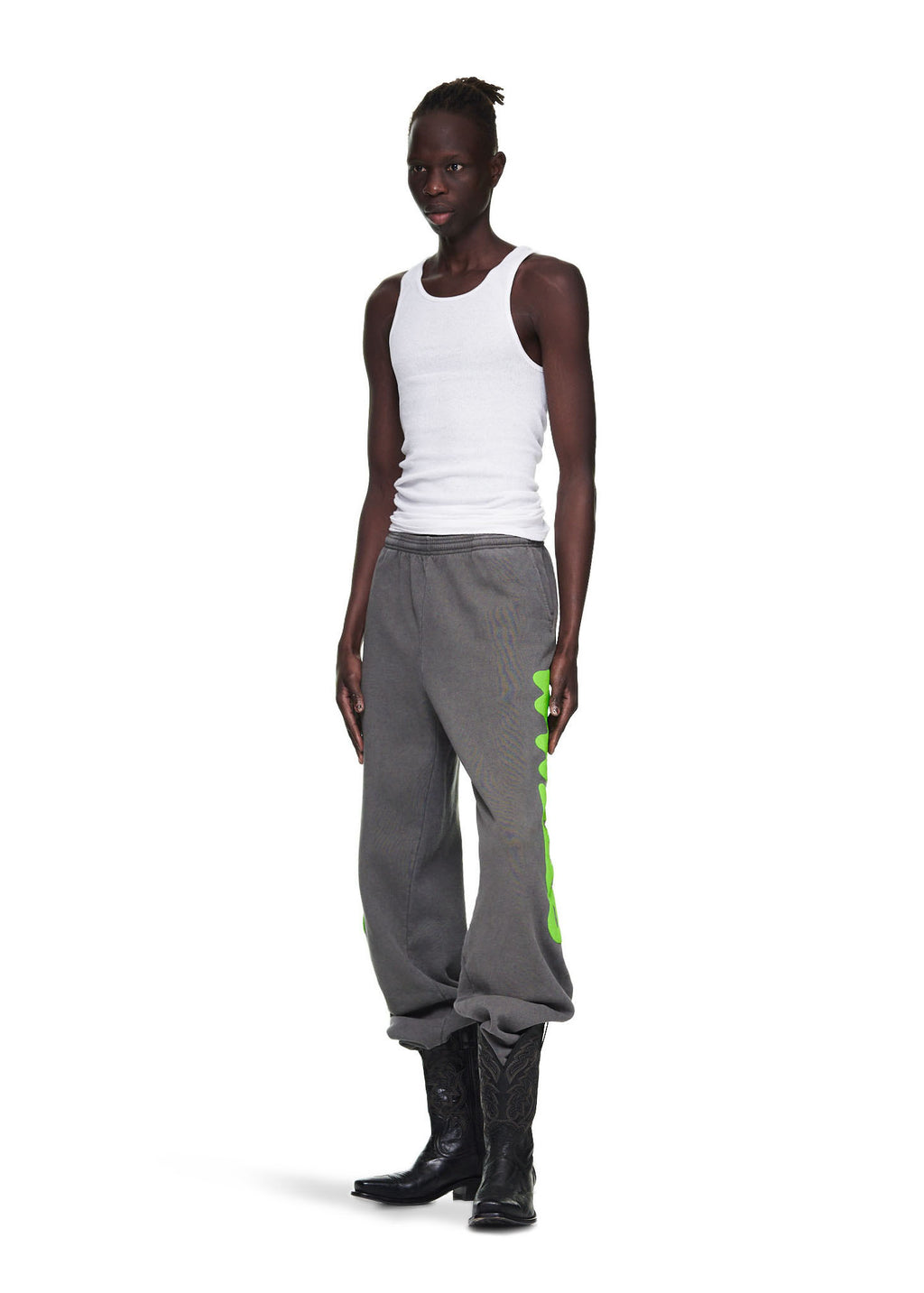 Male model wearing slate grey Beluga sweatpants with neon green 'Worldwide' puff print, paired with a white tank top and black cowboy boots.