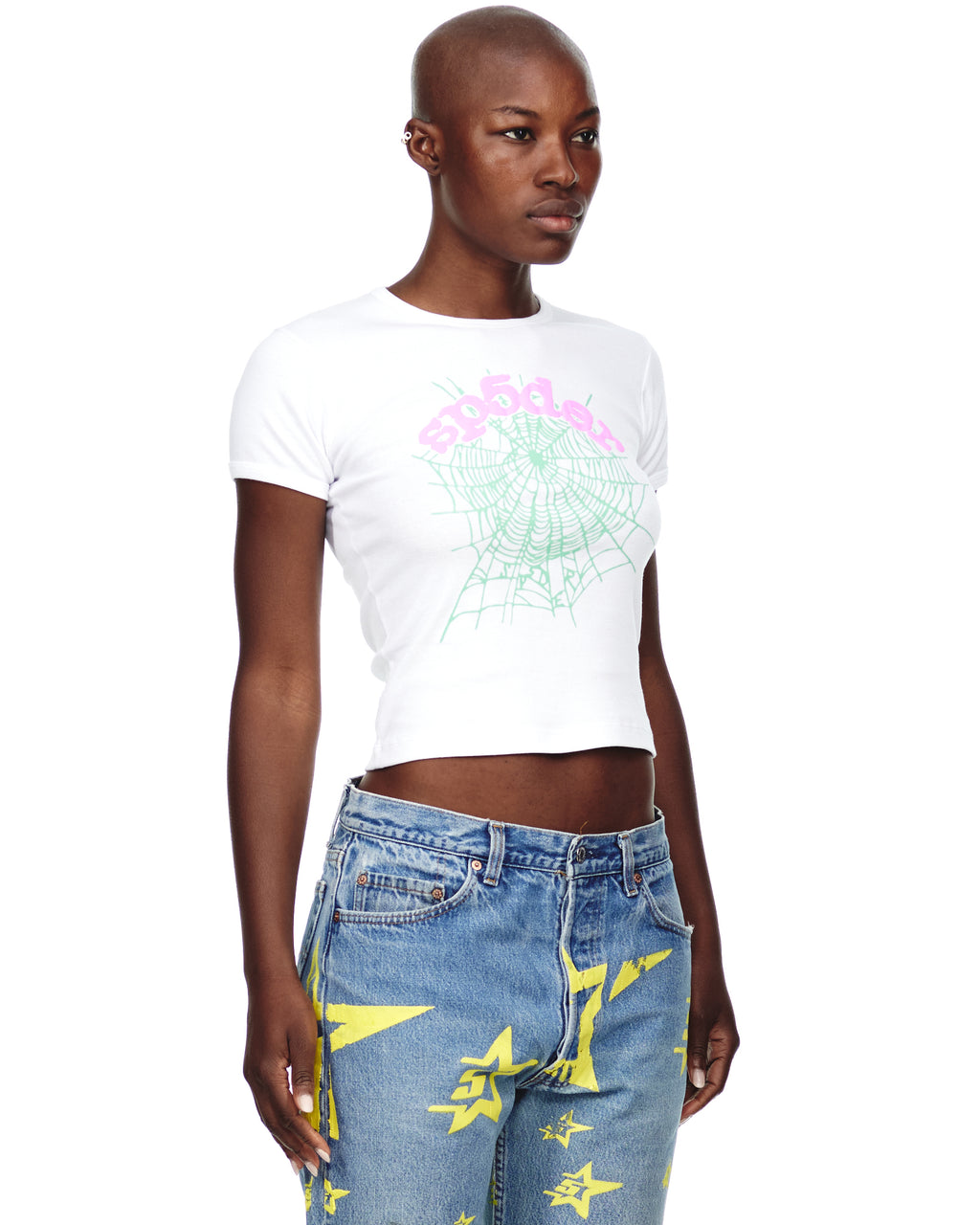 Female model in white OG WEB CANDY BABY TEE with pink text and green spiderweb graphic, paired with blue jeans.