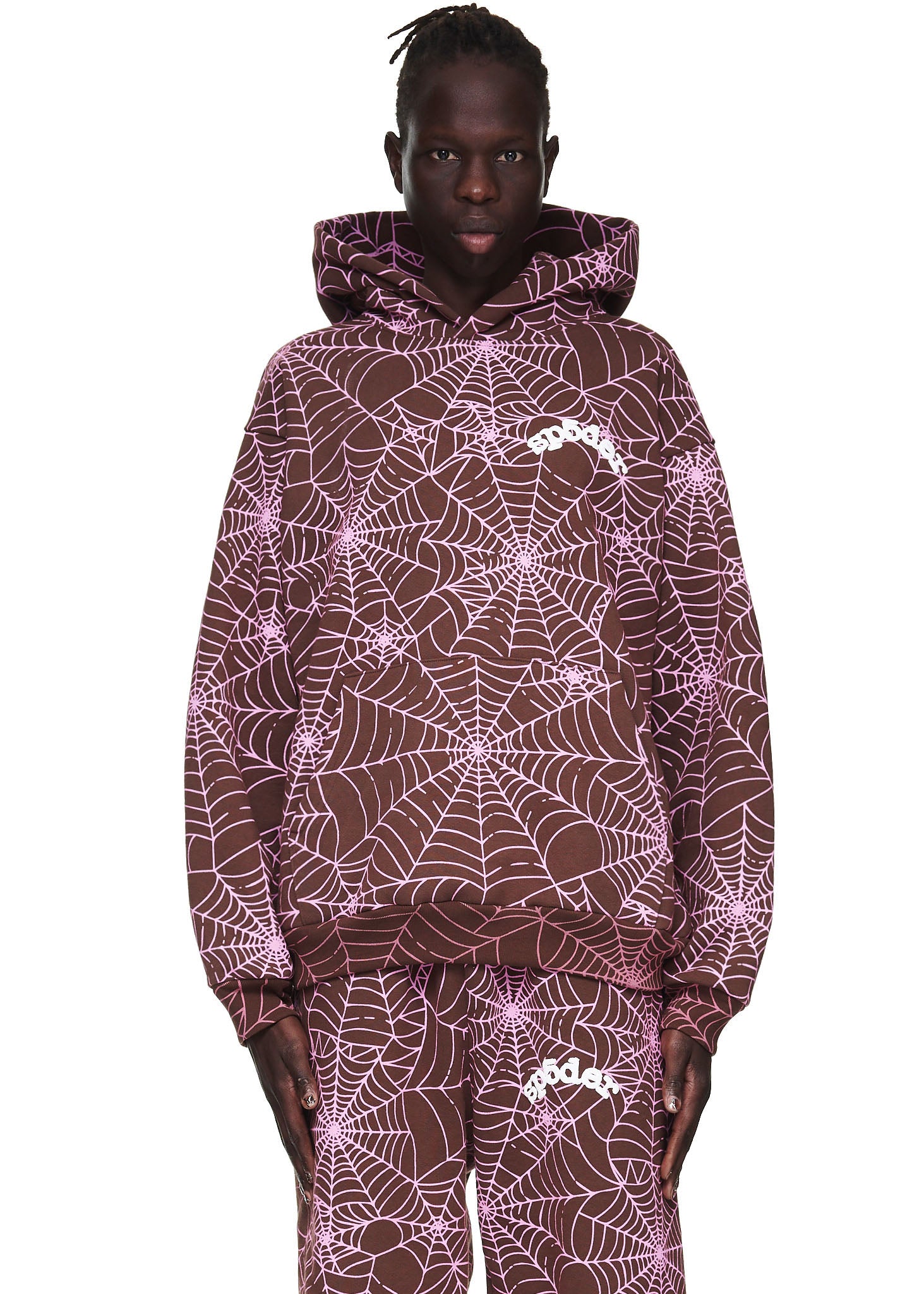 Male model in a brown AOP hoodie with pink cracked web print and "SP5DER" text.