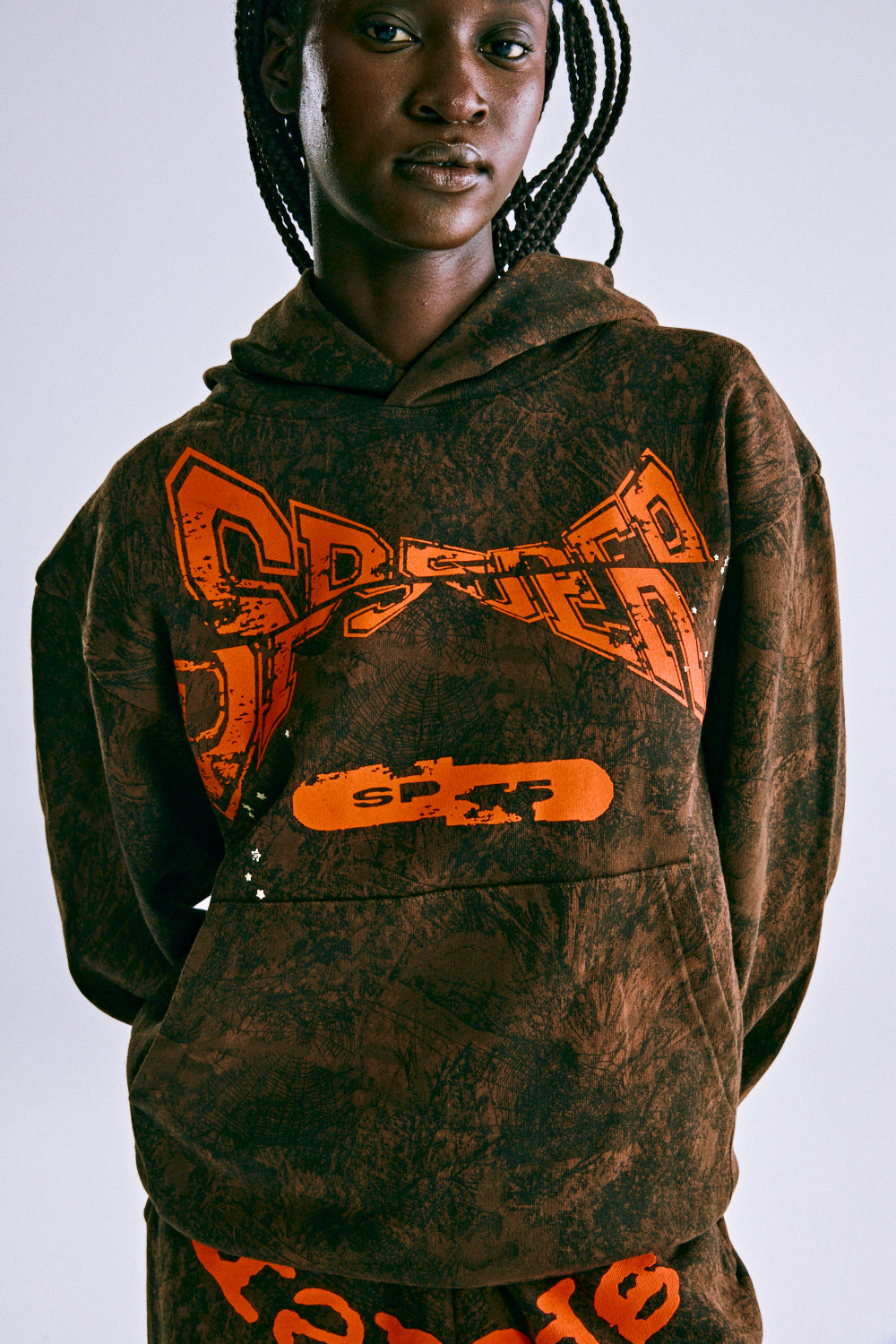 Brown tree camo hoodie with orange "SP5DER" chest print, kangaroo pocket, and rib knit details.