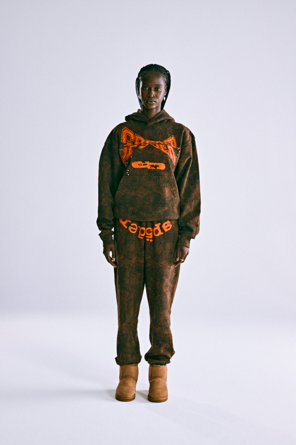 Brown tree camo hoodie and sweatpants set with orange "SP5DER" text, worn by a model.
