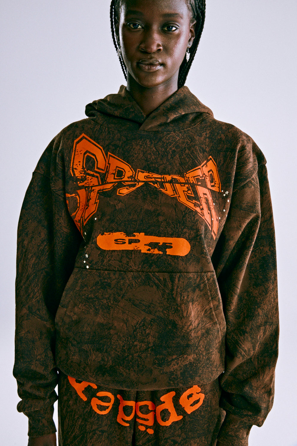 Brown tree camo hoodie with orange puff print "SP5DER" text, kangaroo pocket, and rib knit details.