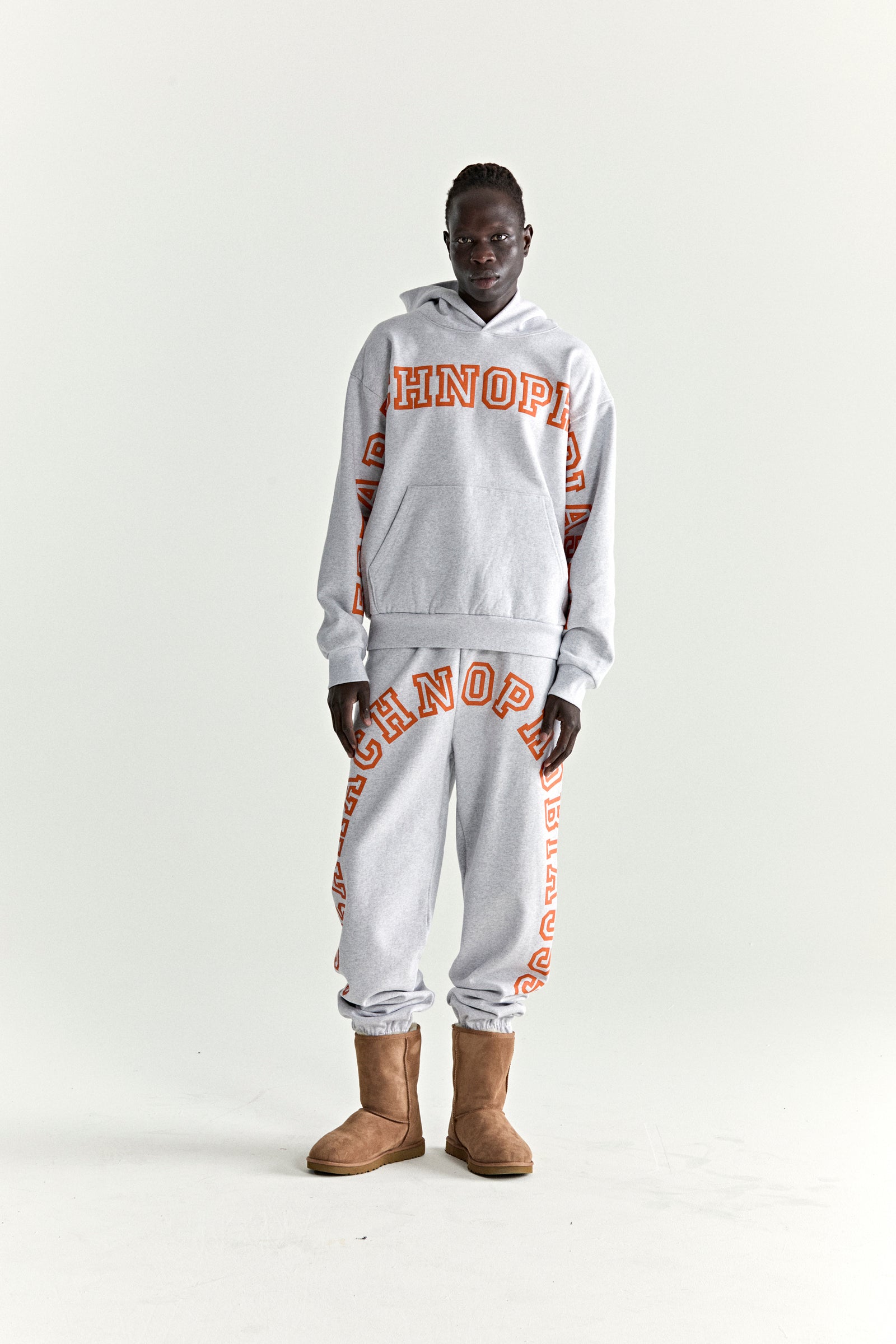 Model wearing a grey ARACH HOODIE and sweatpants with orange puff print text, kangaroo pocket, and rib knit details.
