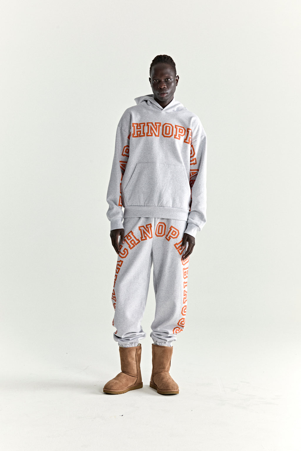 Model wearing a grey ARACH HOODIE and sweatpants with orange puff print text, kangaroo pocket, and rib knit details.