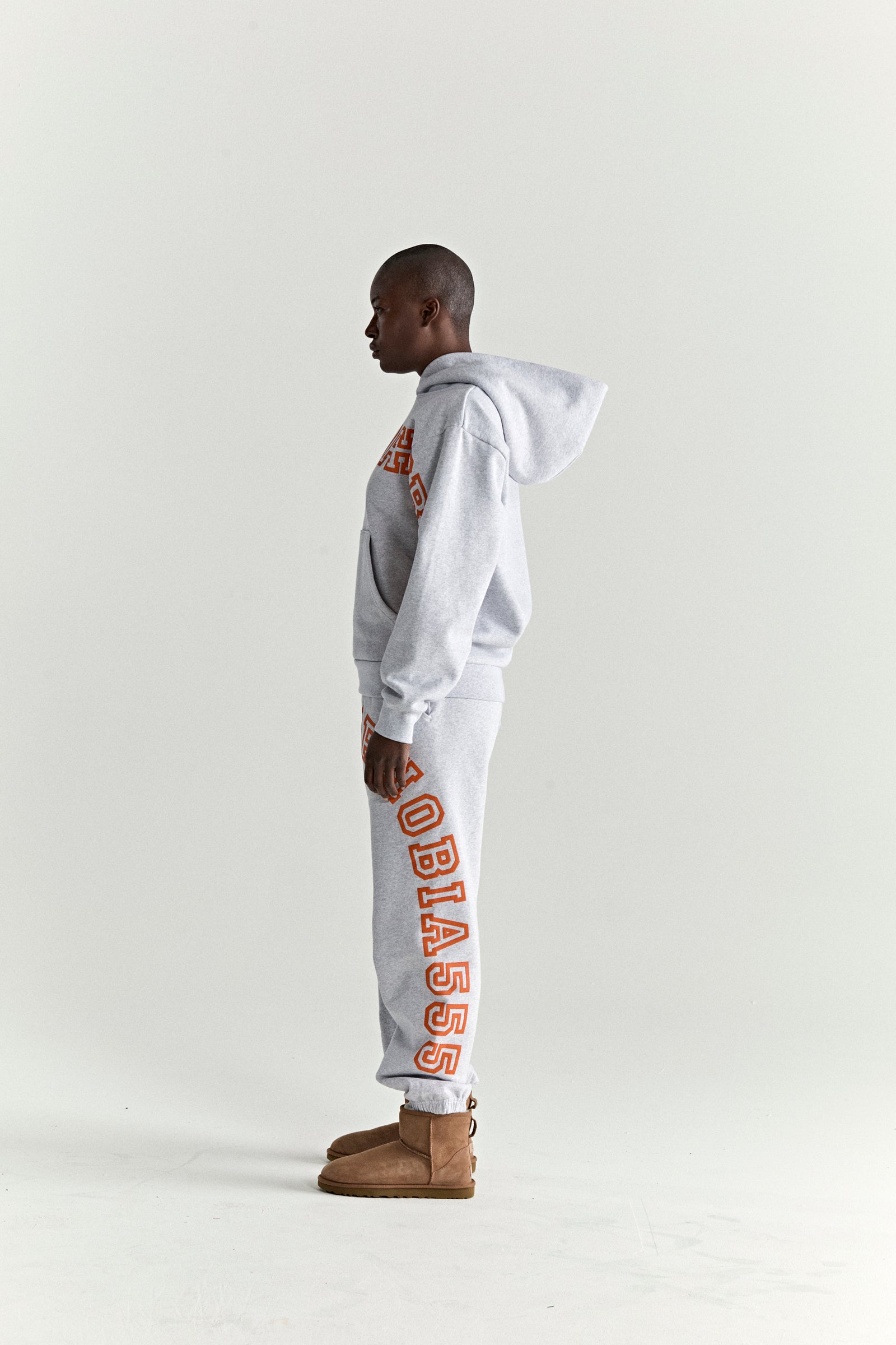 Grey 13oz cotton fleece hoodie and sweatpants with orange puff print text, worn by a model in profile.