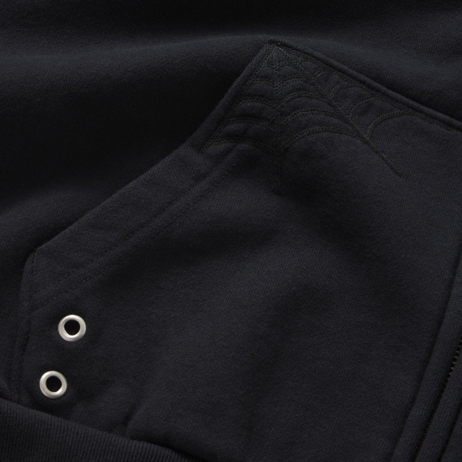 Black 13oz cotton fleece zip hoodie featuring tonal web embroidery on the pocket and silver metal grommet detailing.