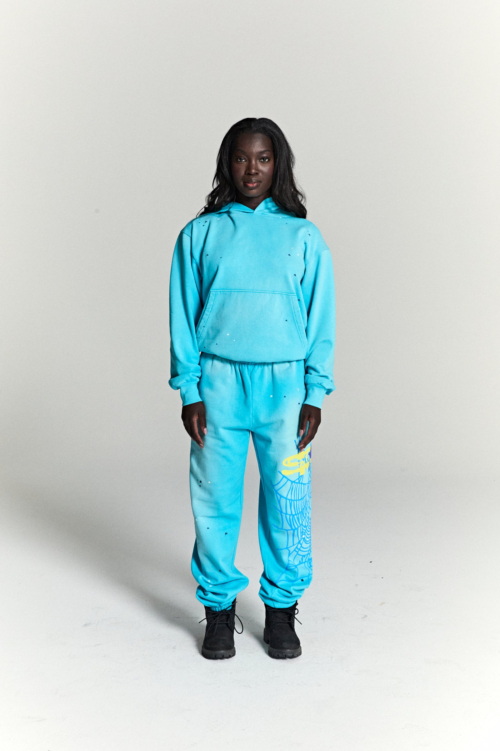 Woman in a turquoise HEAVY WEB HOODIE and sweatpants with scattered stars and a yellow spider web print.