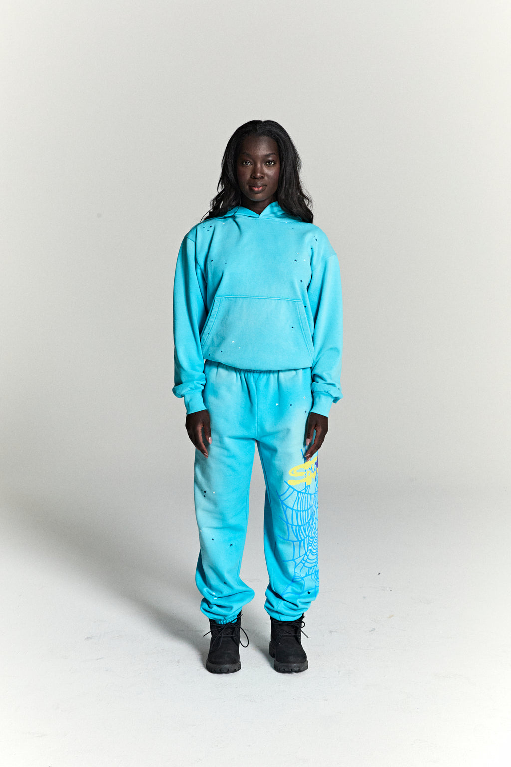 Woman in a turquoise HEAVY WEB HOODIE and sweatpants with scattered stars and a yellow spider web print.
