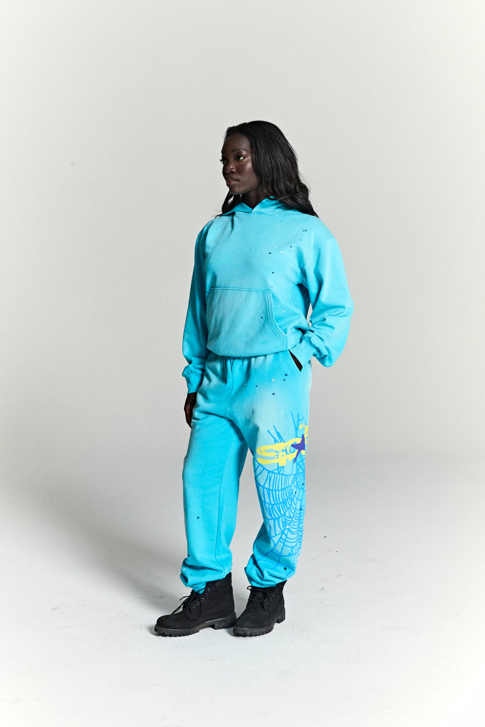 Woman in a turquoise HEAVY WEB HOODIE and matching sweatpants with a yellow web print.