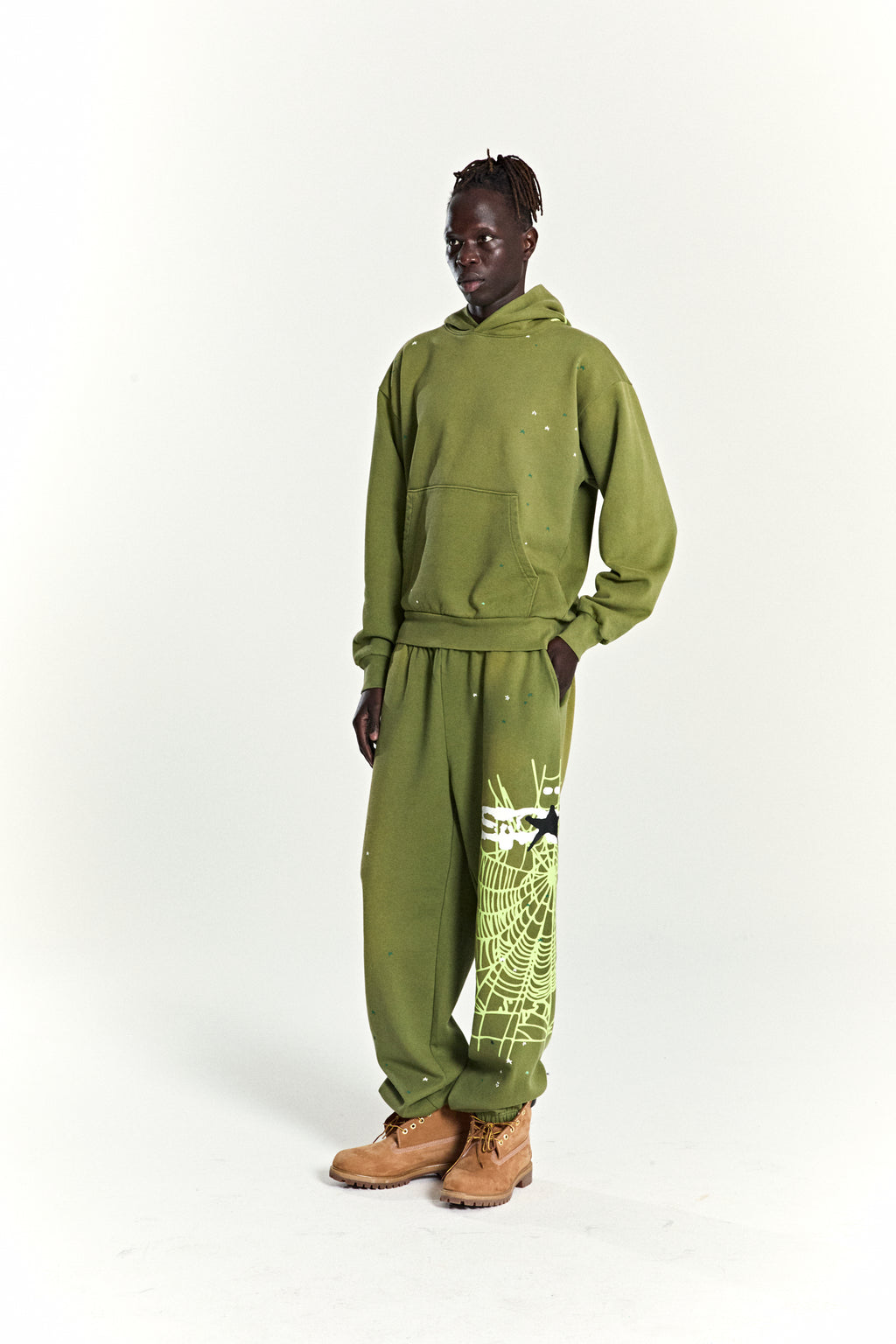 Model in olive green HEAVY WEB HOODIE and sweatpants with scattered stars and a large web print.