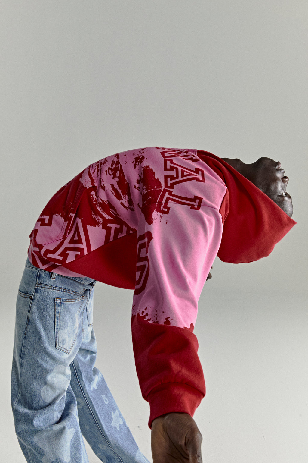 NOCTURNAL ZIP HOODIE: Red and pink color-blocked 13oz cotton fleece hoodie with varsity lettering, worn by a person in light wash jeans.
