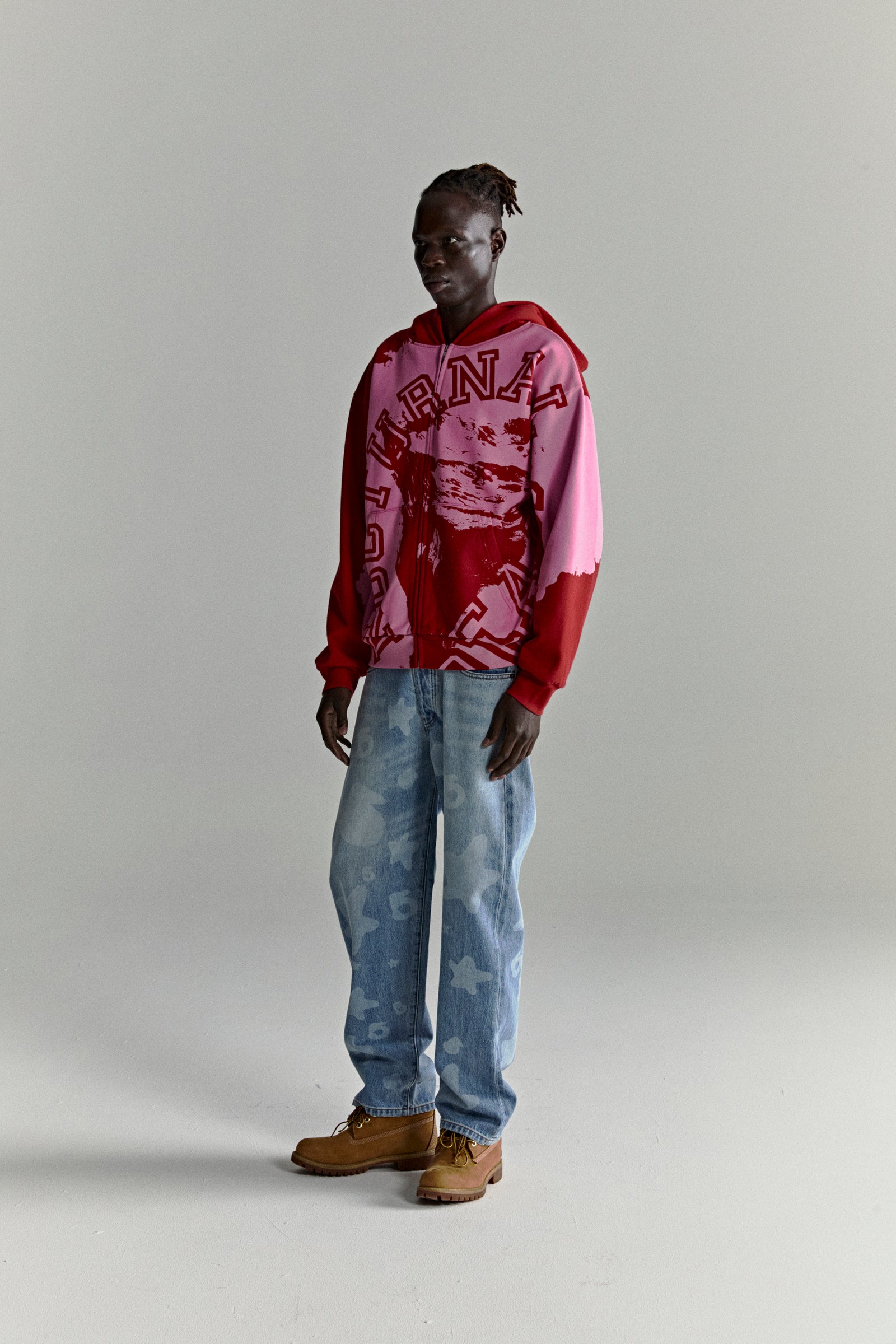 Model wearing a color-blocked red and pink NOCTURNAL ZIP HOODIE with varsity lettering and light wash jeans.