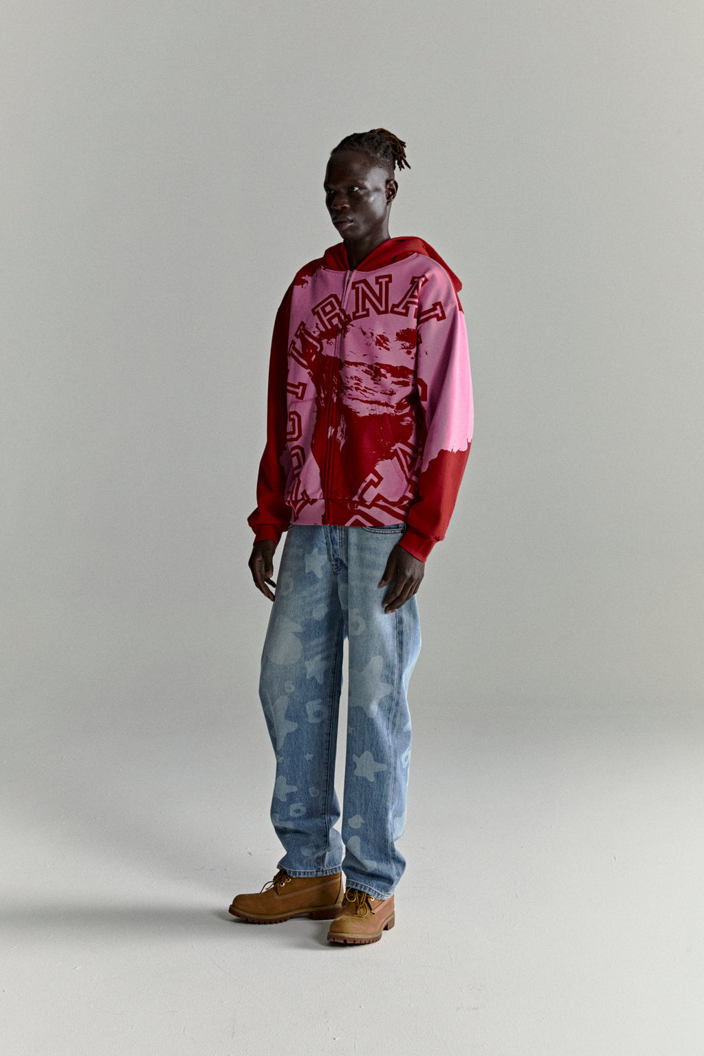 Model wearing a color-blocked red and pink NOCTURNAL ZIP HOODIE with varsity lettering and light wash jeans.