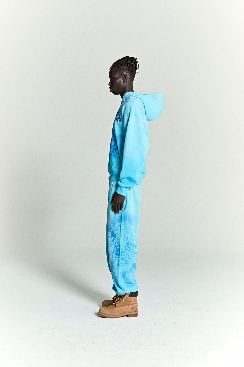 Light blue cotton fleece hoodie and sweatpants with spray web print, worn by a model in profile.