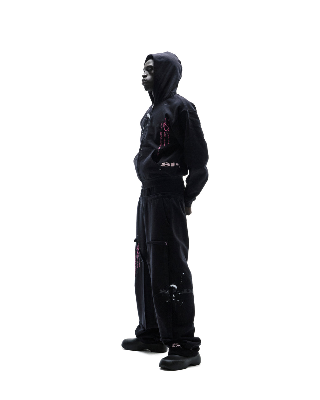Model wearing a black zip-up hoodie and matching baggy sweatpants with pink graphic details against a white background.