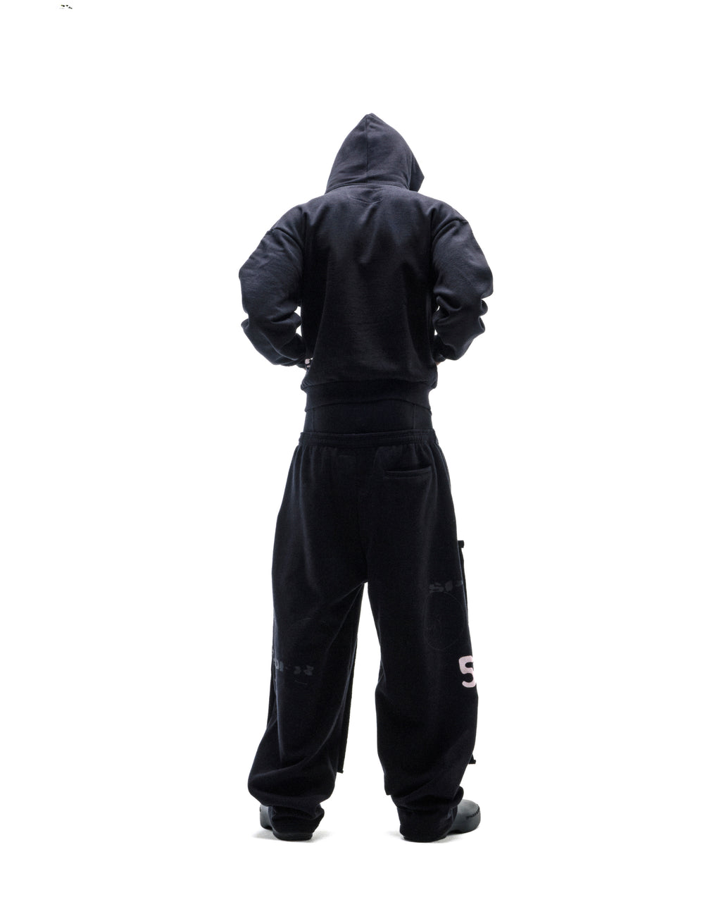 Model wearing a black hooded sweatshirt and oversized black sweatpants, viewed from behind against a white background.