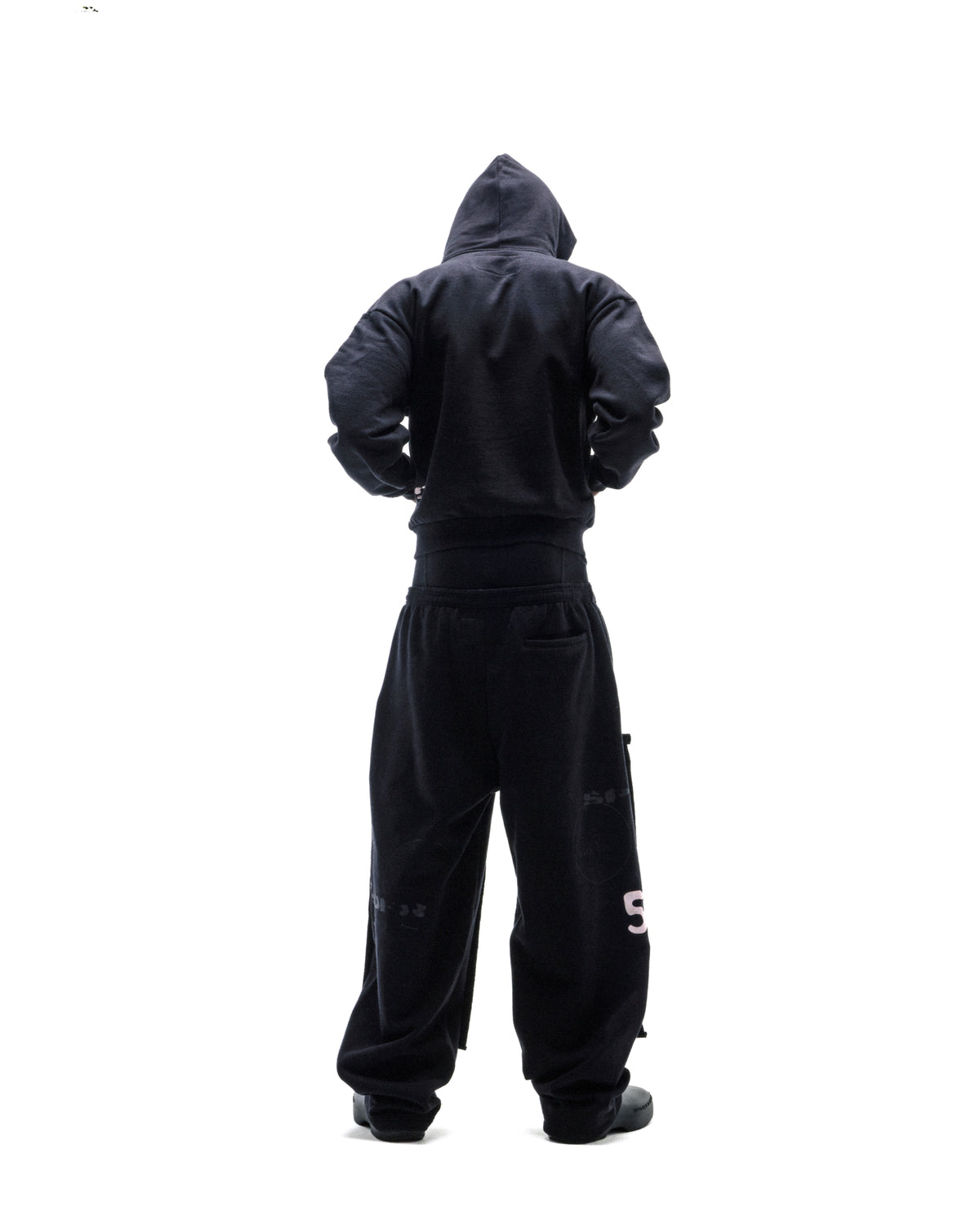 Model wearing a black hooded sweatshirt and oversized black sweatpants, viewed from behind against a white background.