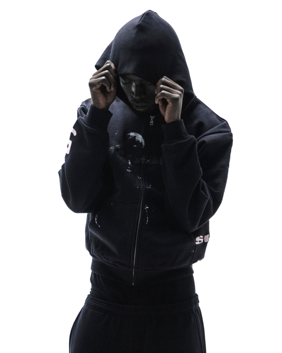 Model wearing a black zip-up hoodie with white graphic details, hood pulled up, isolated against a white background.