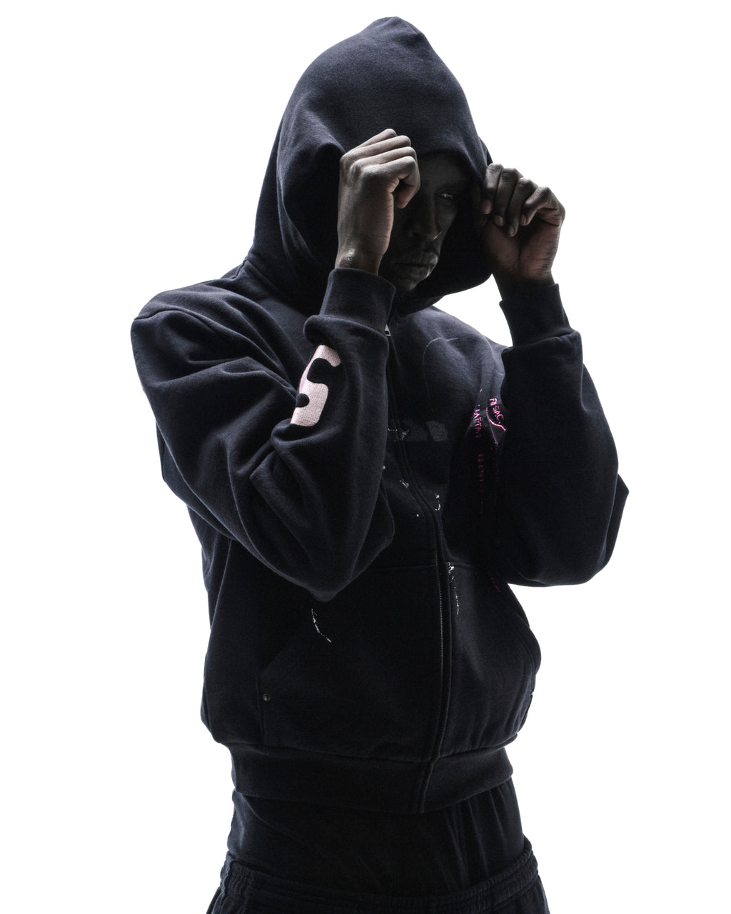 Model wearing a black zip-up hoodie with pink accents and a hood pulled up against a bright white background.