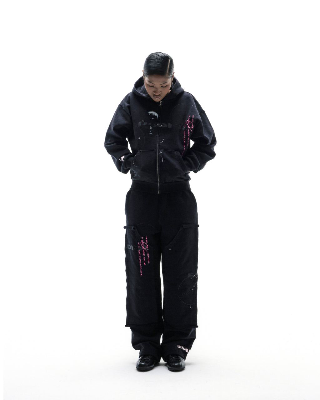 Model wearing a black zip-up hoodie and matching cargo pants with pink graphic details against a white background.