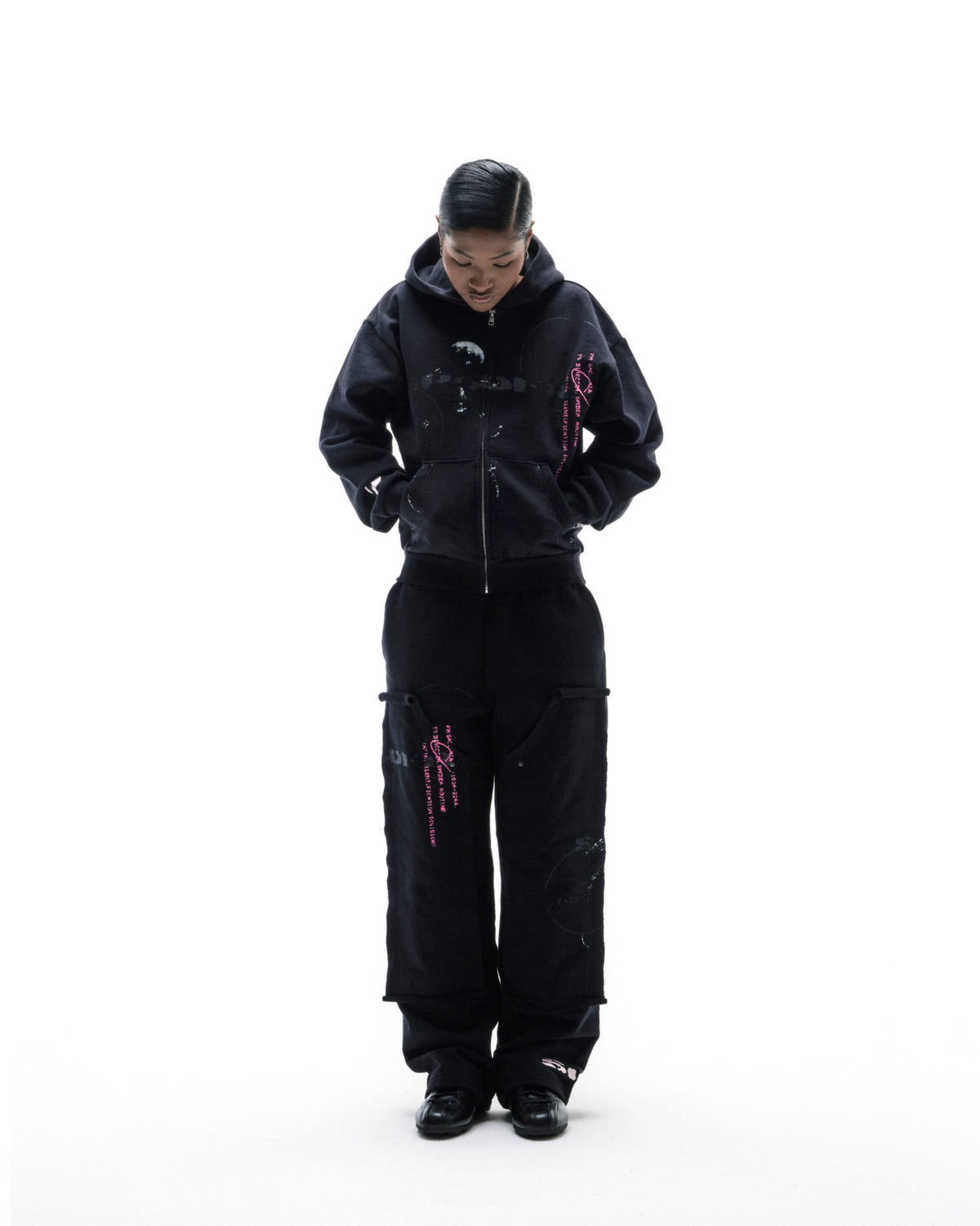Model wearing a black zip-up hoodie and matching cargo pants with pink graphic details against a white background.
