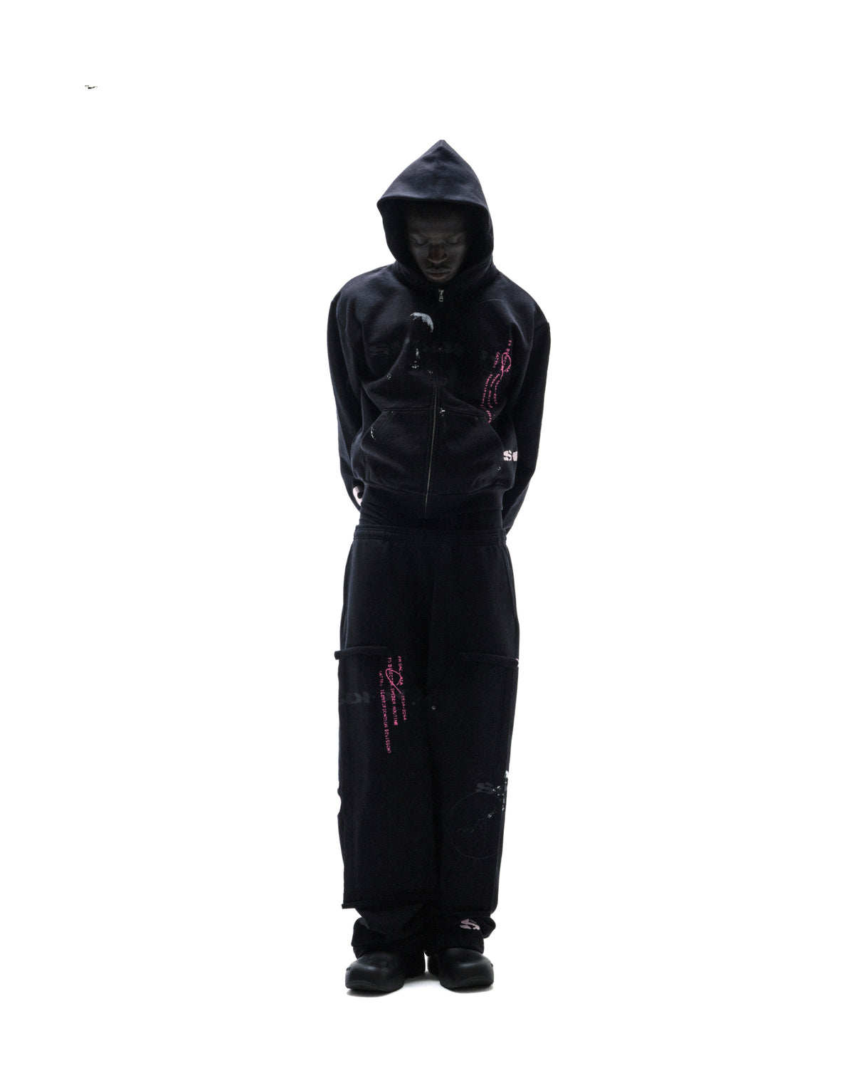 Model wearing a black zip-up hoodie and matching sweatpants with pink graphic prints against a white background.