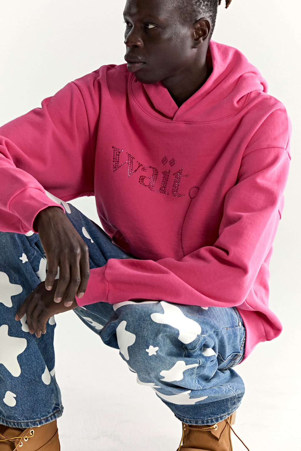 Model wearing a pink cotton fleece hoodie with rhinestone "Wait" detail and blue jeans.