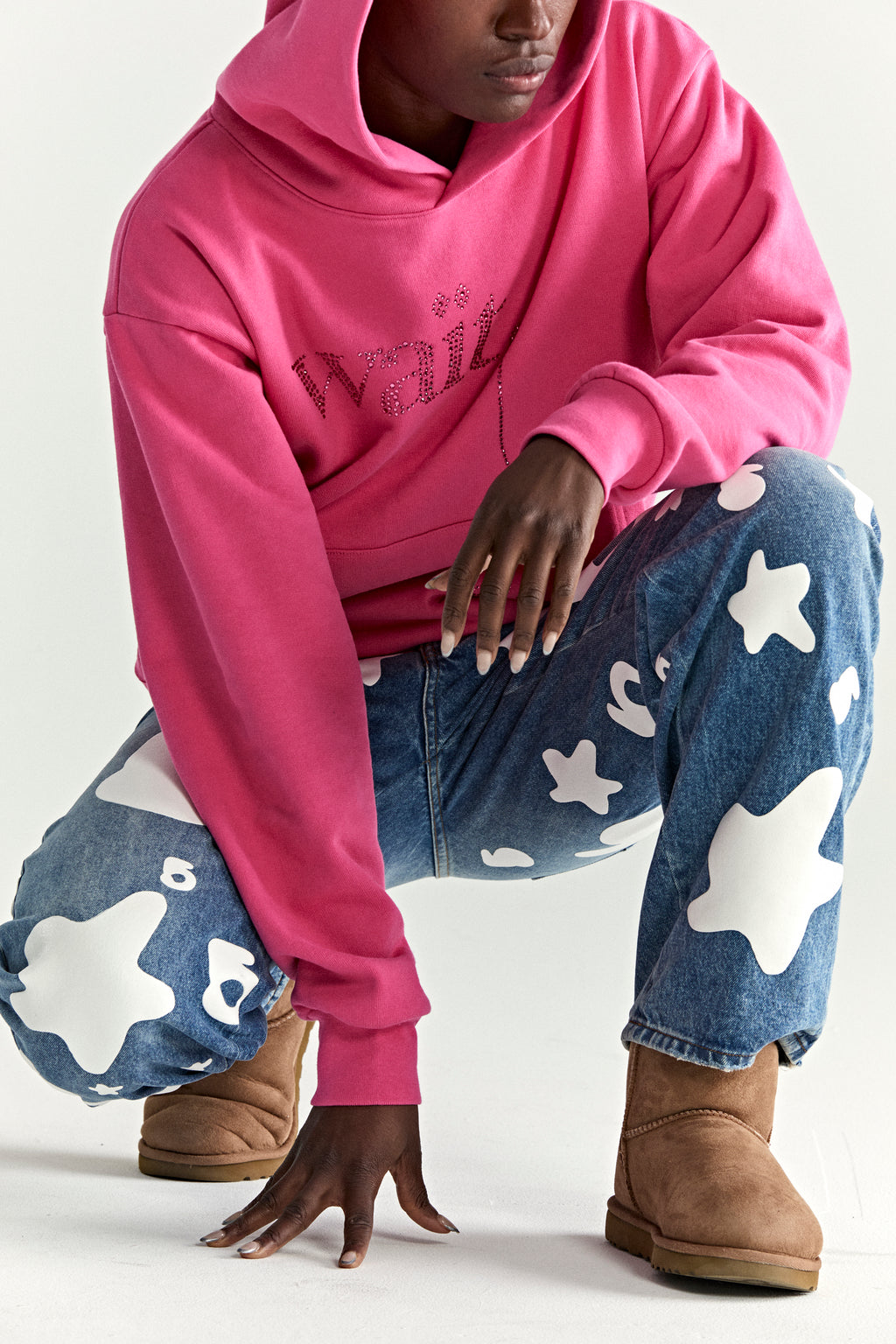 Pink cotton fleece hoodie with rhinestone "Wait" detail, worn with patterned jeans and brown boots.