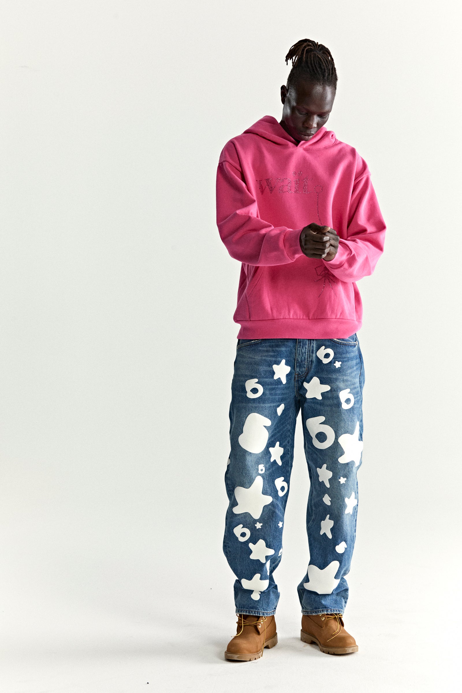Model in a pink Wait Stone Hoodie with rhinestone detail, blue jeans with white star and number patterns, and brown boots.