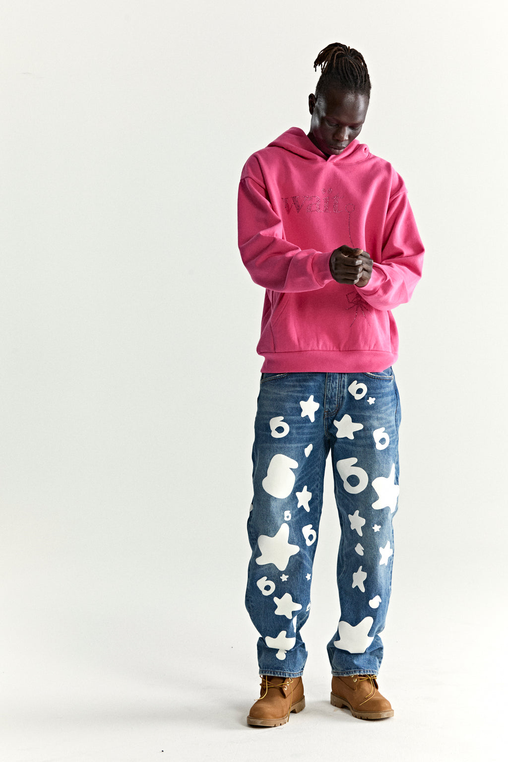 Model in a pink Wait Stone Hoodie with rhinestone detail, blue jeans with white star and number patterns, and brown boots.