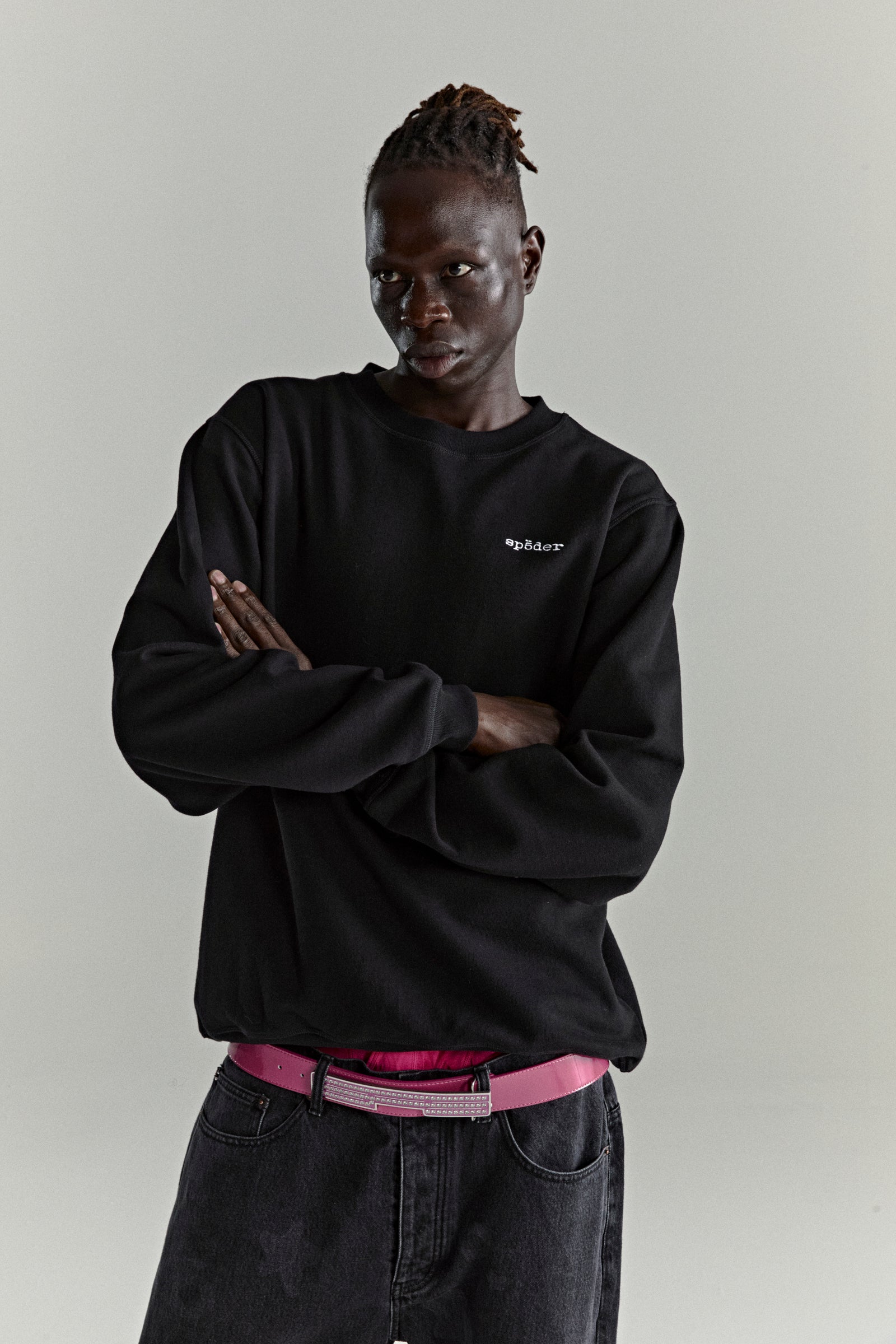 Man in black CORE CREWNECK with embroidered 'SP5DER' logo, dark jeans, and pink belt.