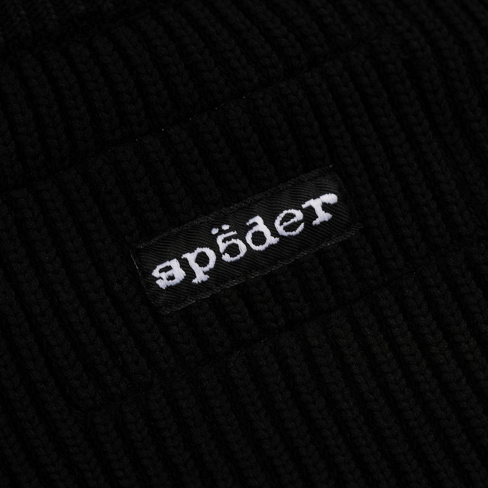 Close-up of black ribbed knit fabric with a black woven label featuring "apöder" in white embroidery.