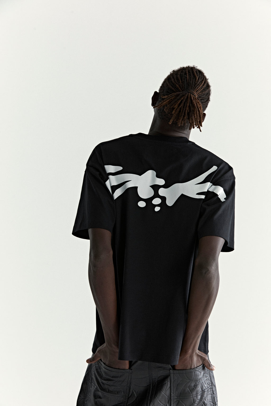 Black 100% cotton TM Tee with a white graphic print across the back, worn by a person with dreadlocks.