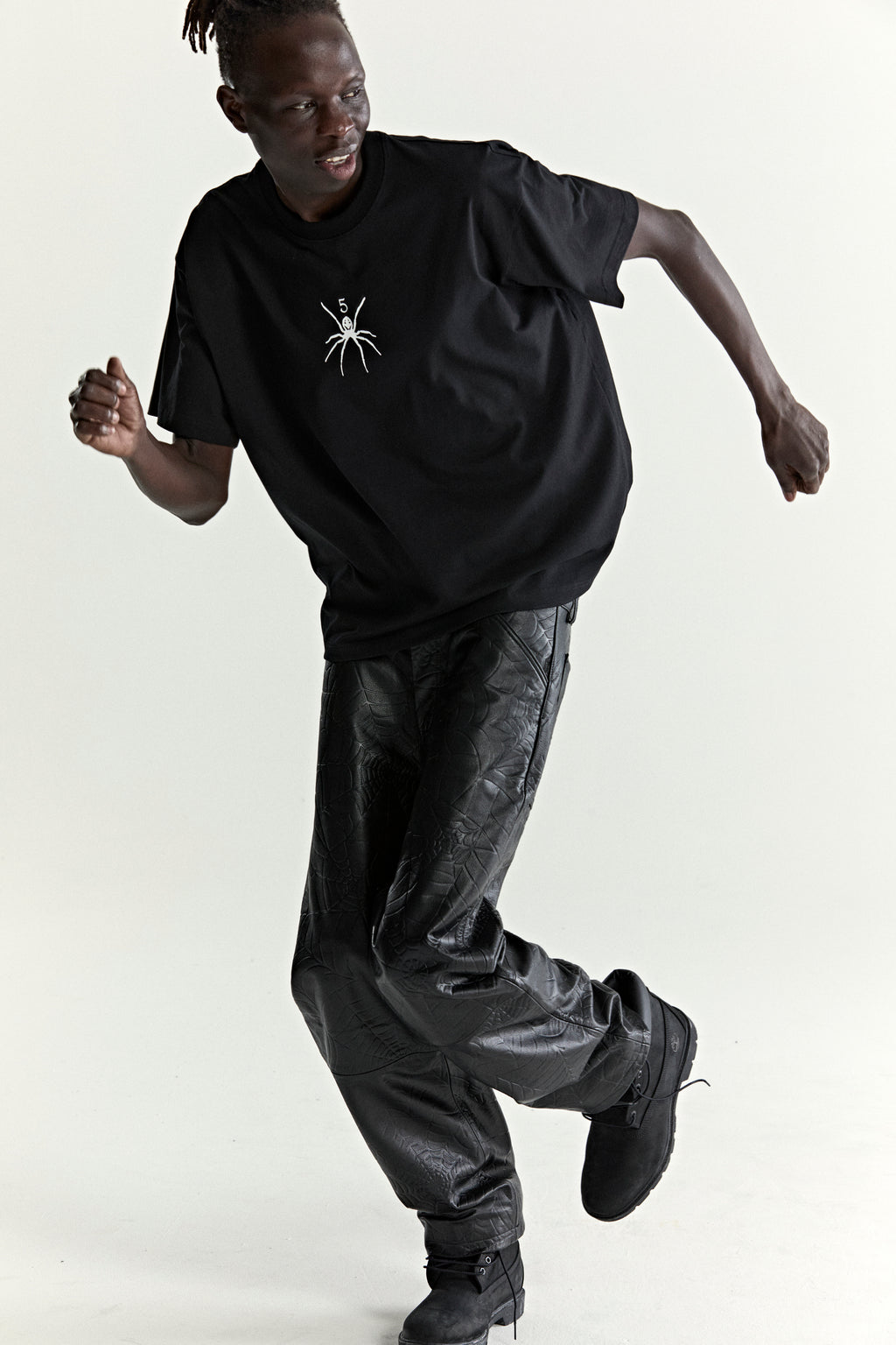 Black 100% cotton TM Tee with white spider graphic, worn by a man in black patterned pants and boots.