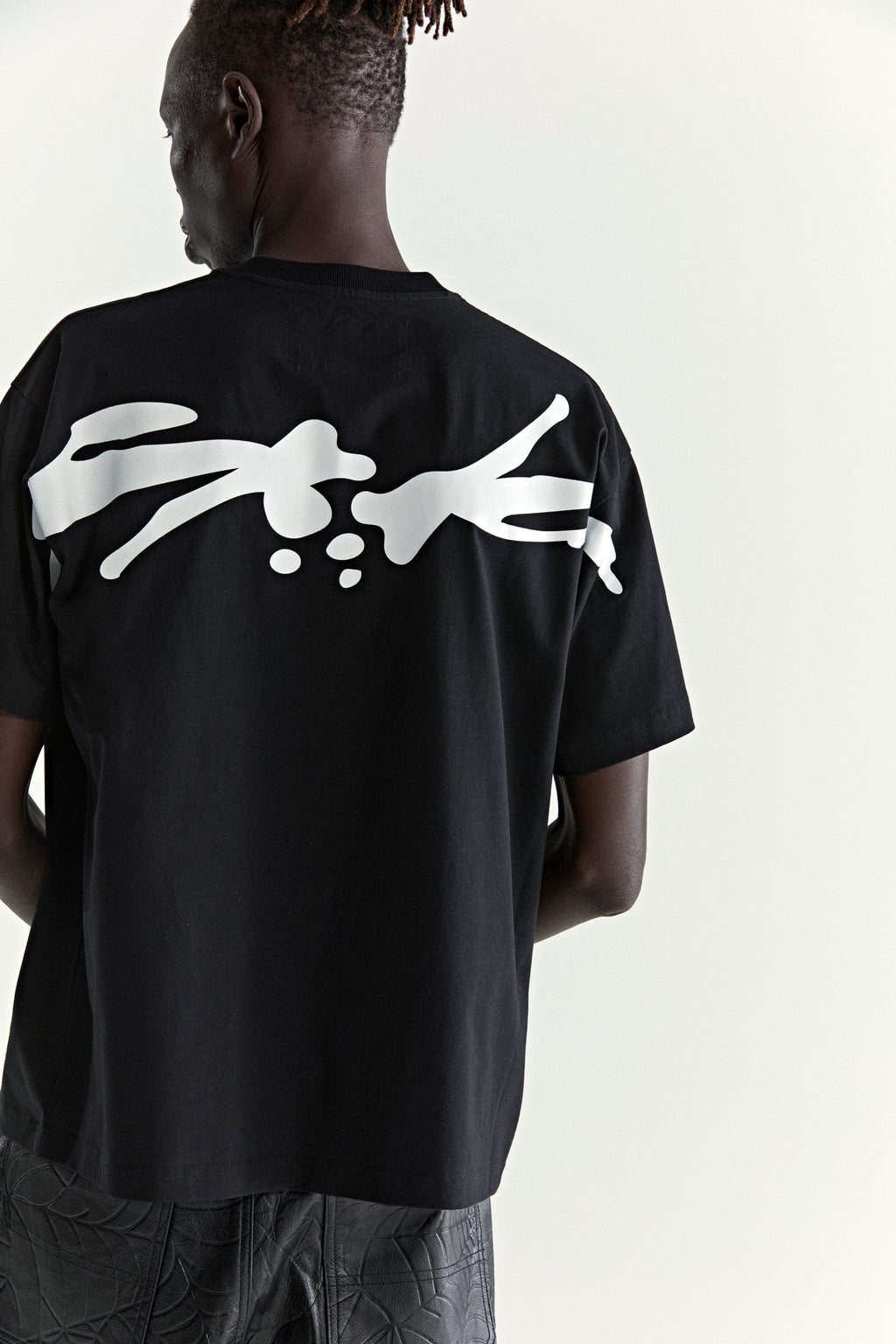 Black 100% cotton TM TEE with a large white graphic print on the back, worn by a person.