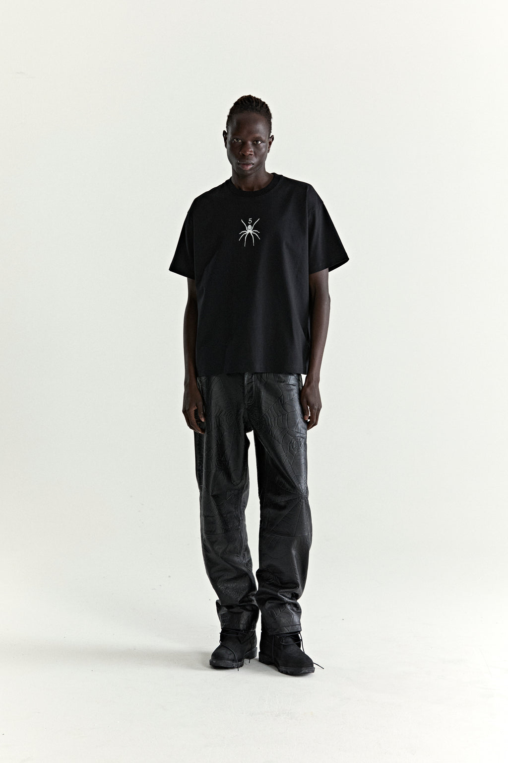 Black 100% cotton TM TEE with white spider graphic, worn by a model with black patterned pants.