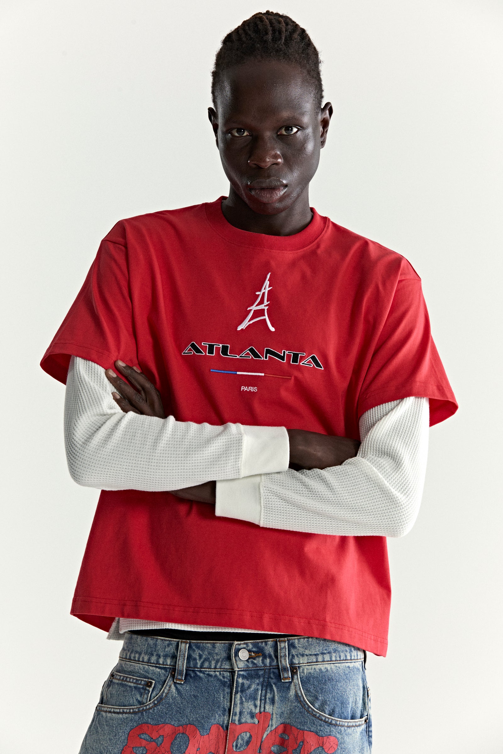 Model in red cotton ARAIGNÉE TEE with embroidered Eiffel Tower and "ATLANTA PARIS" graphic, layered over a white long-sleeve.