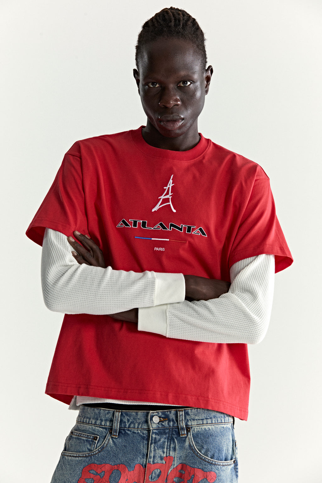 Model in red cotton ARAIGNÉE TEE with embroidered Eiffel Tower and "ATLANTA PARIS" graphic, layered over a white long-sleeve.